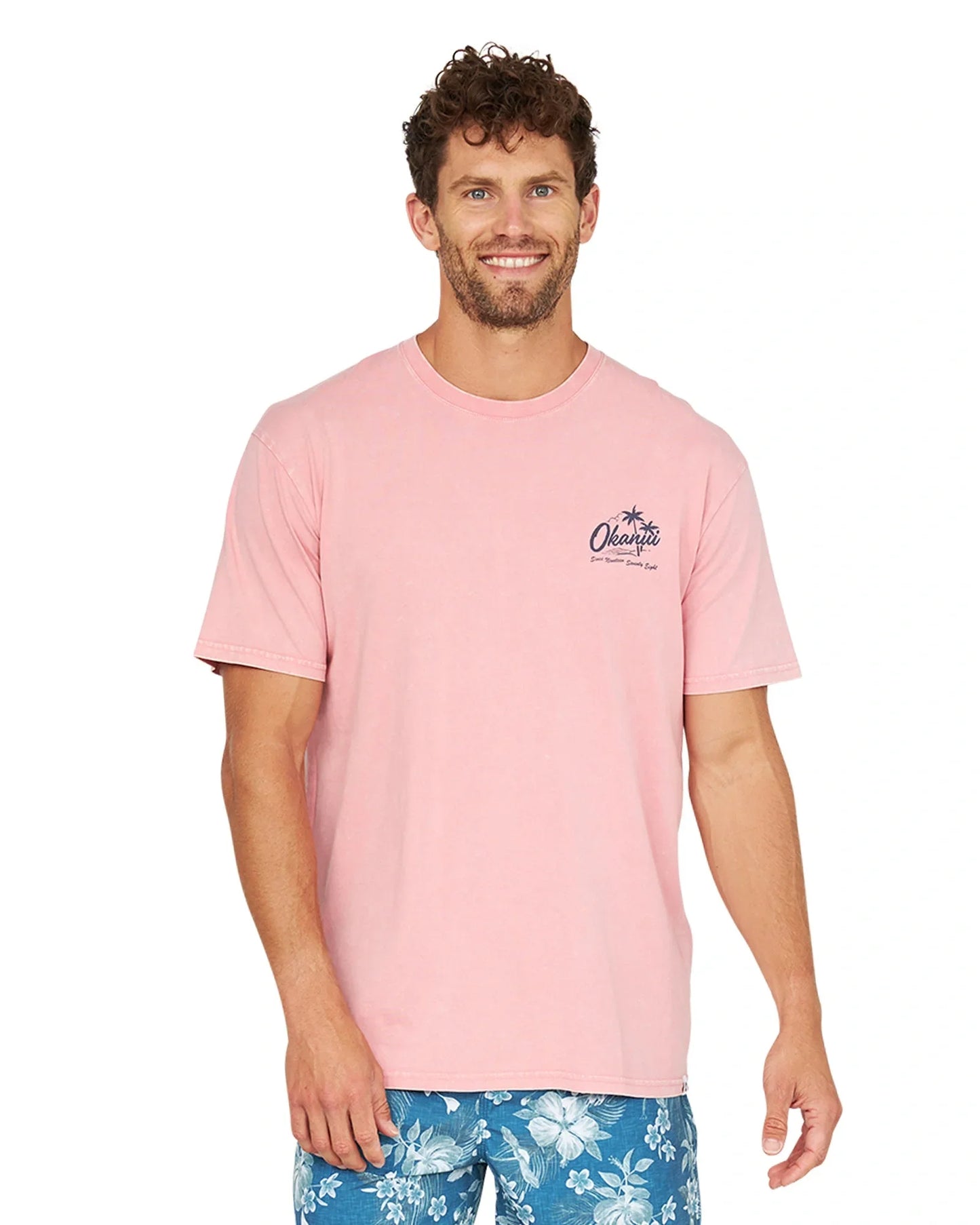 A front view of a model wearing the Okanui Postcards Tee in washed pink color and blue swimshorts.
