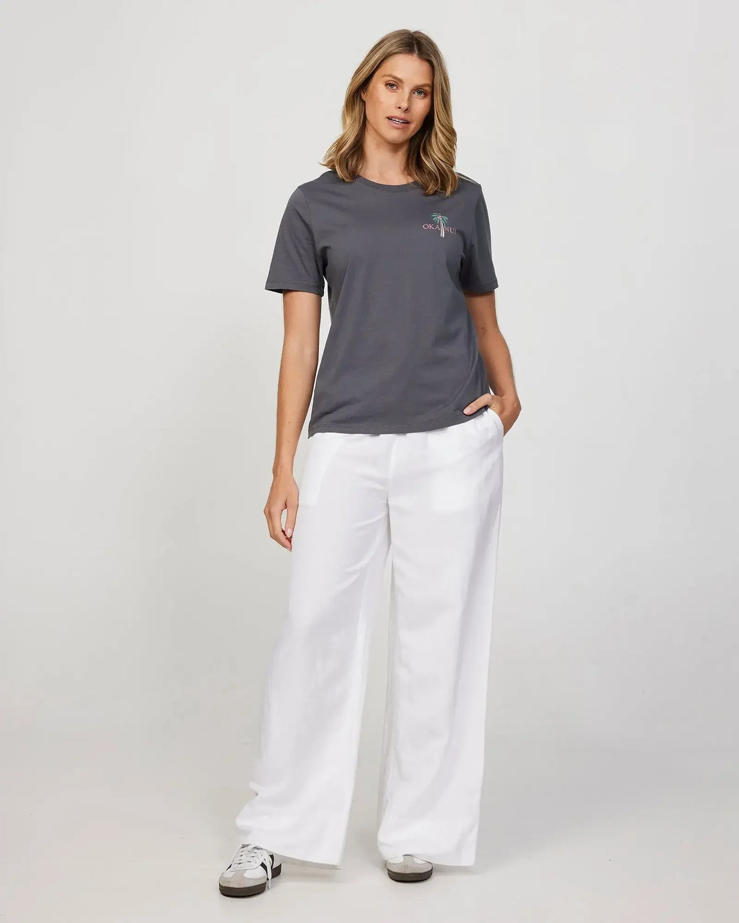 Female model wearing white loose pants and grey Okanui Resort Tee with hand in her pocket.