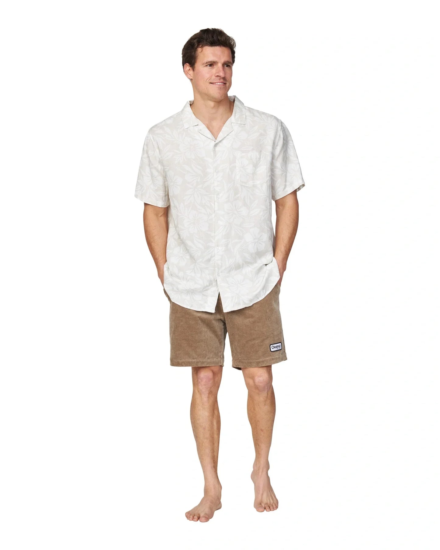 A casual shot of a male model standing with hands in pockets wearing the Okanui Sandblast Short Sleeve Aloha Shirt in stone colour.