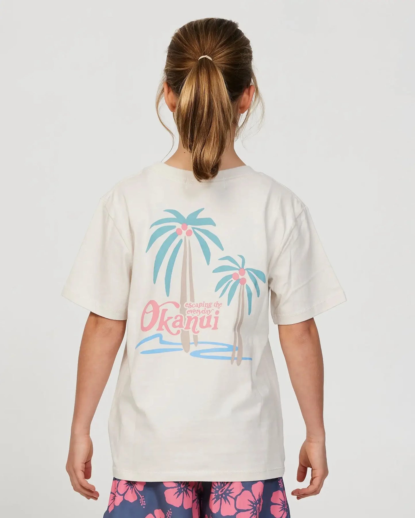 Back view of a little girl model wearing the Okanui Twin Palm Tee T-shirt in Stone