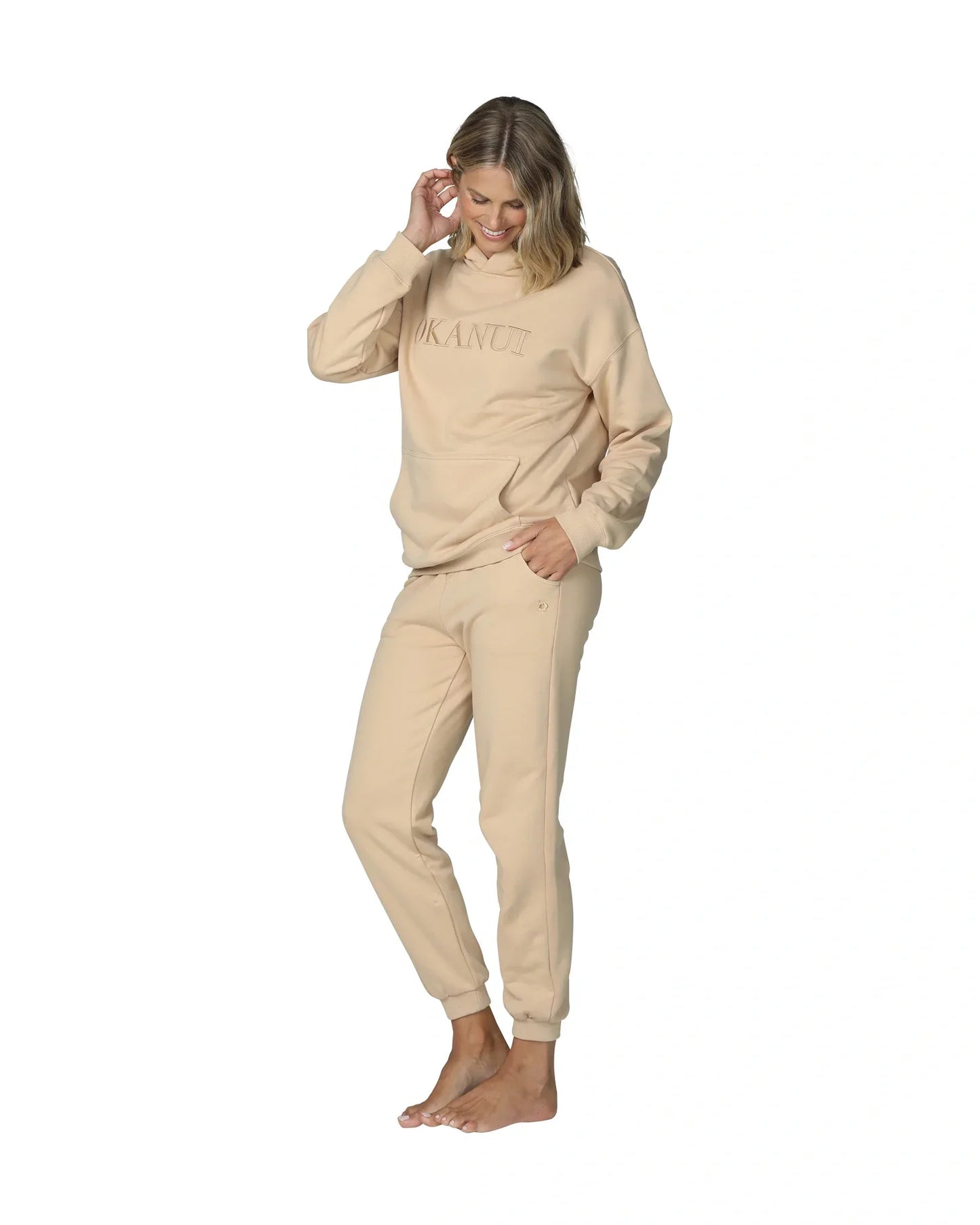 Full body front view of a female model with her hand in her hair, wearing a natural color jacket paired with the Okanui jogger track pants in natural color