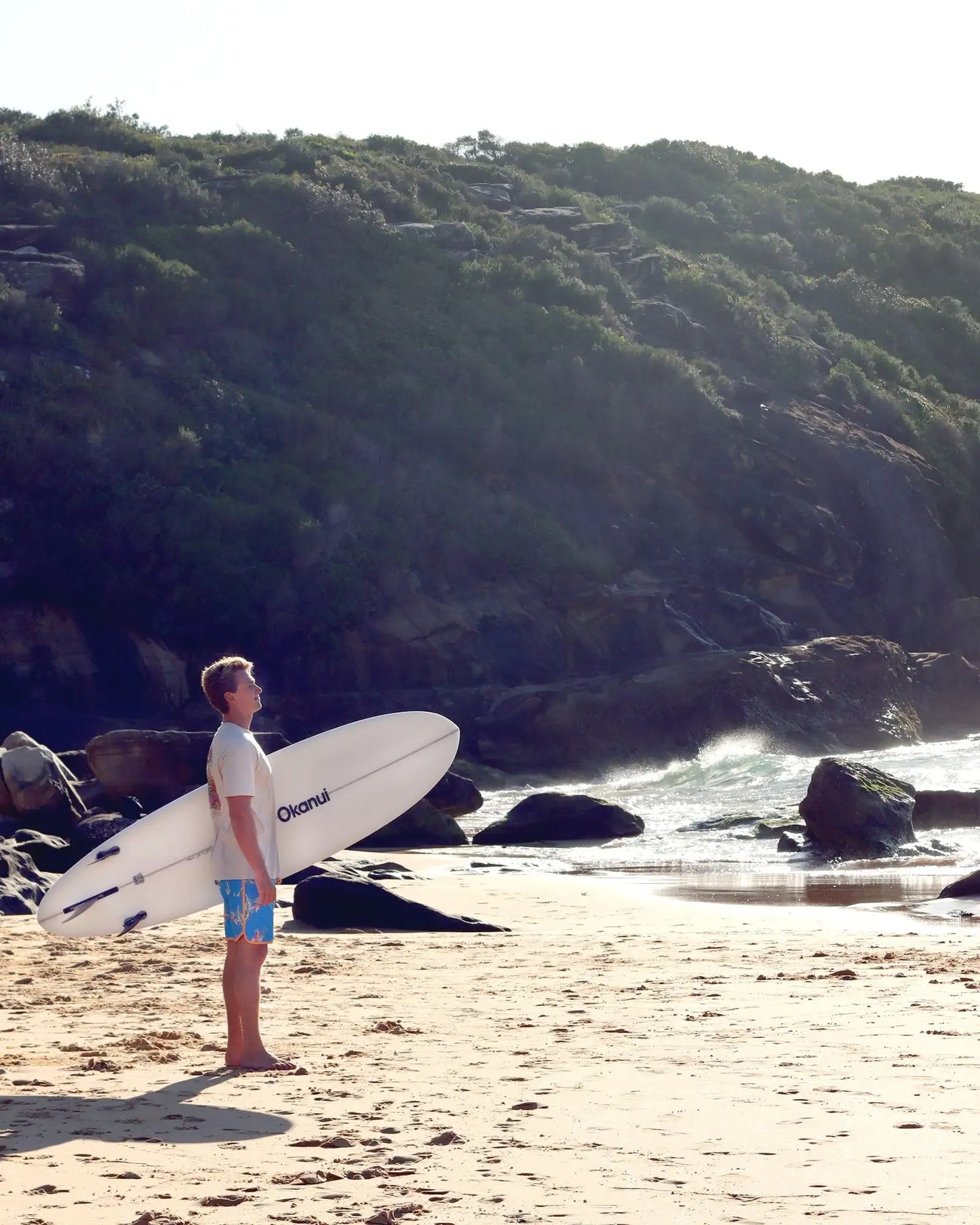 A male person holding the White Okanui The Bucket Mid Length Surfboard with Okanui logo print