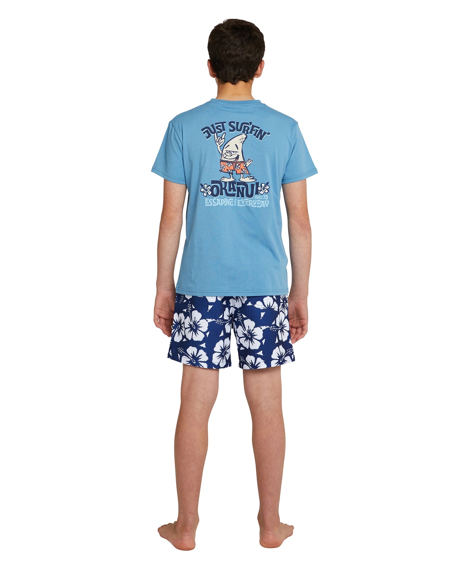 Full body back view of a boy wearing the Okanui boys swim shorts in Hibiscus Navy paired with a t-shirt