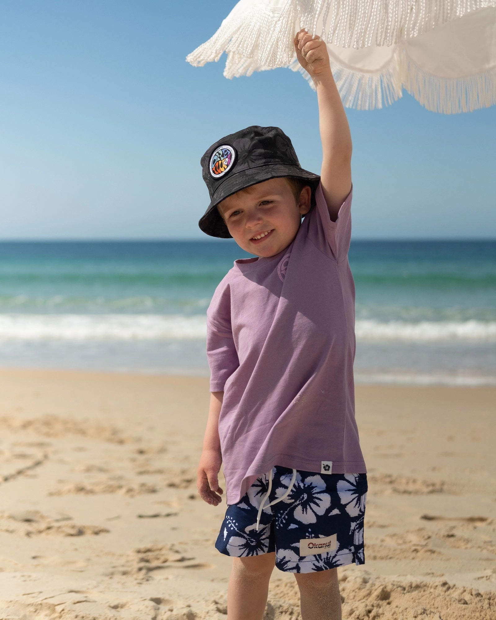 A baby boy in a bucket hat and t-shirt wearing the Okanui boys swim shorts in Hibiscus Navy