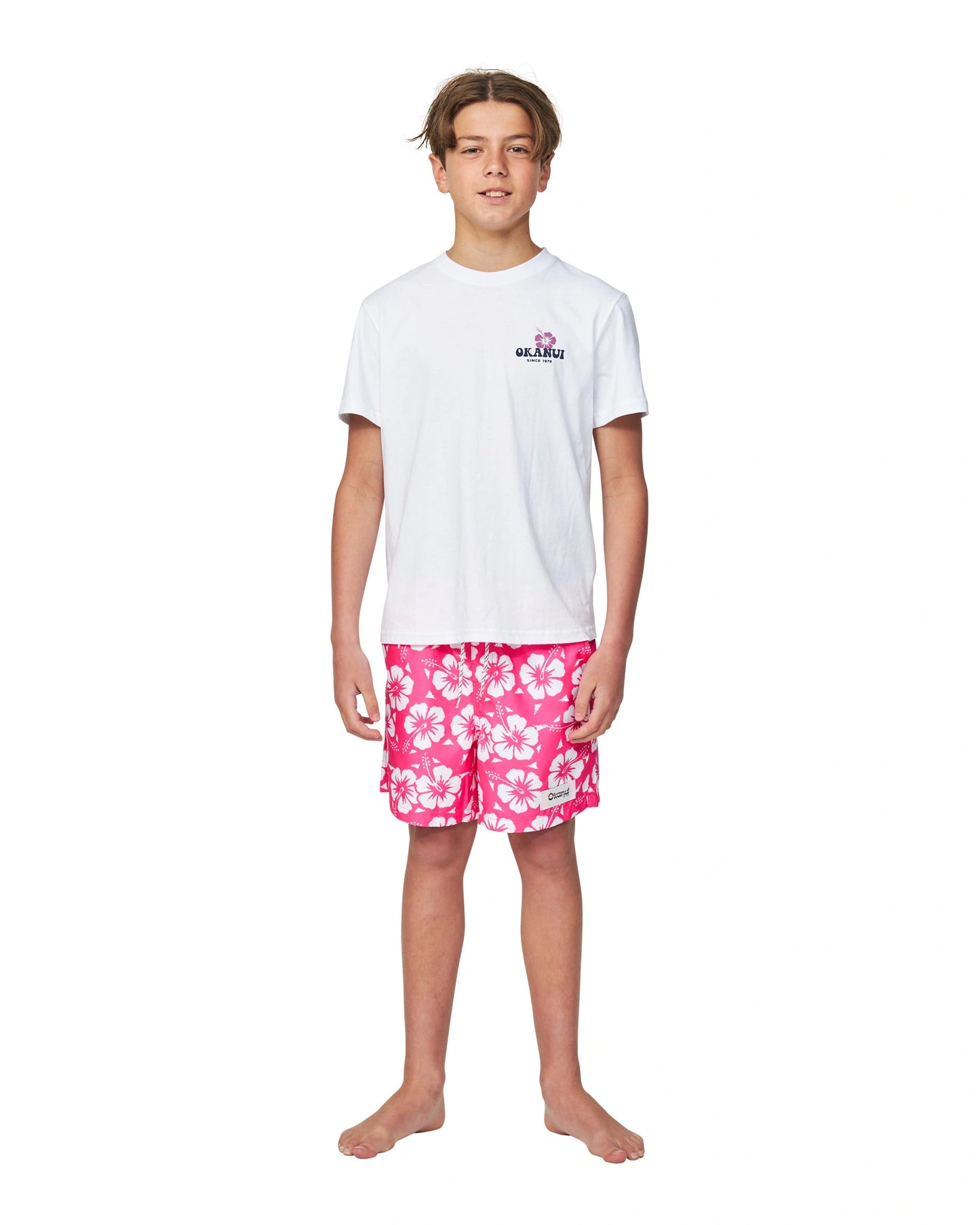 Full body front view of a boy wearing the Okanui boys swim shorts in Hibiscus Hot Pink paired with a white t-shirt