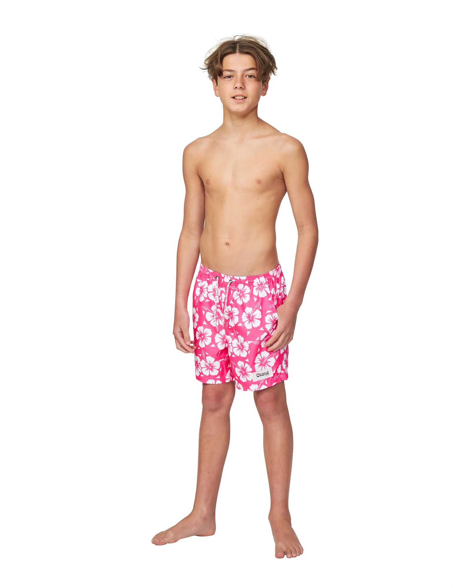 A topless boy wearing the Okanui boys swim shorts in Hibiscus Hot Pink