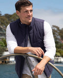 A close up view of male model leaning on the bow of a yatch wearing the Okanui Navy Heather Port Vest in Navy colour over a white long sleeve shirt.
