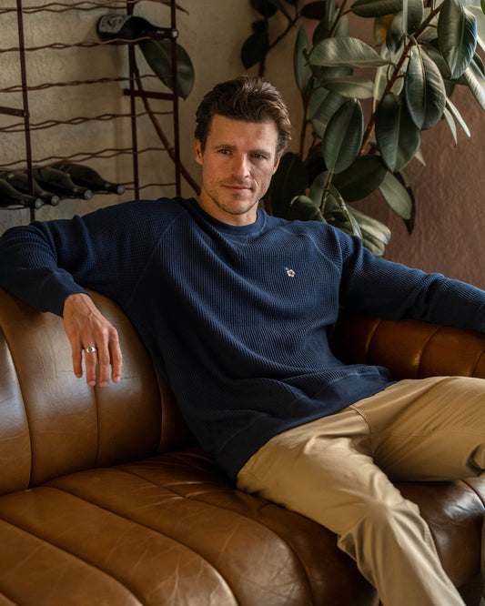 A sitting male model wearing the Okanui Long Sleeve waffle knit jumper in Navy