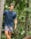 A male model posing with trees and plants in the background wearing the Okanui Noosa Retro Boardrider Boardshort in Flint Stone color.