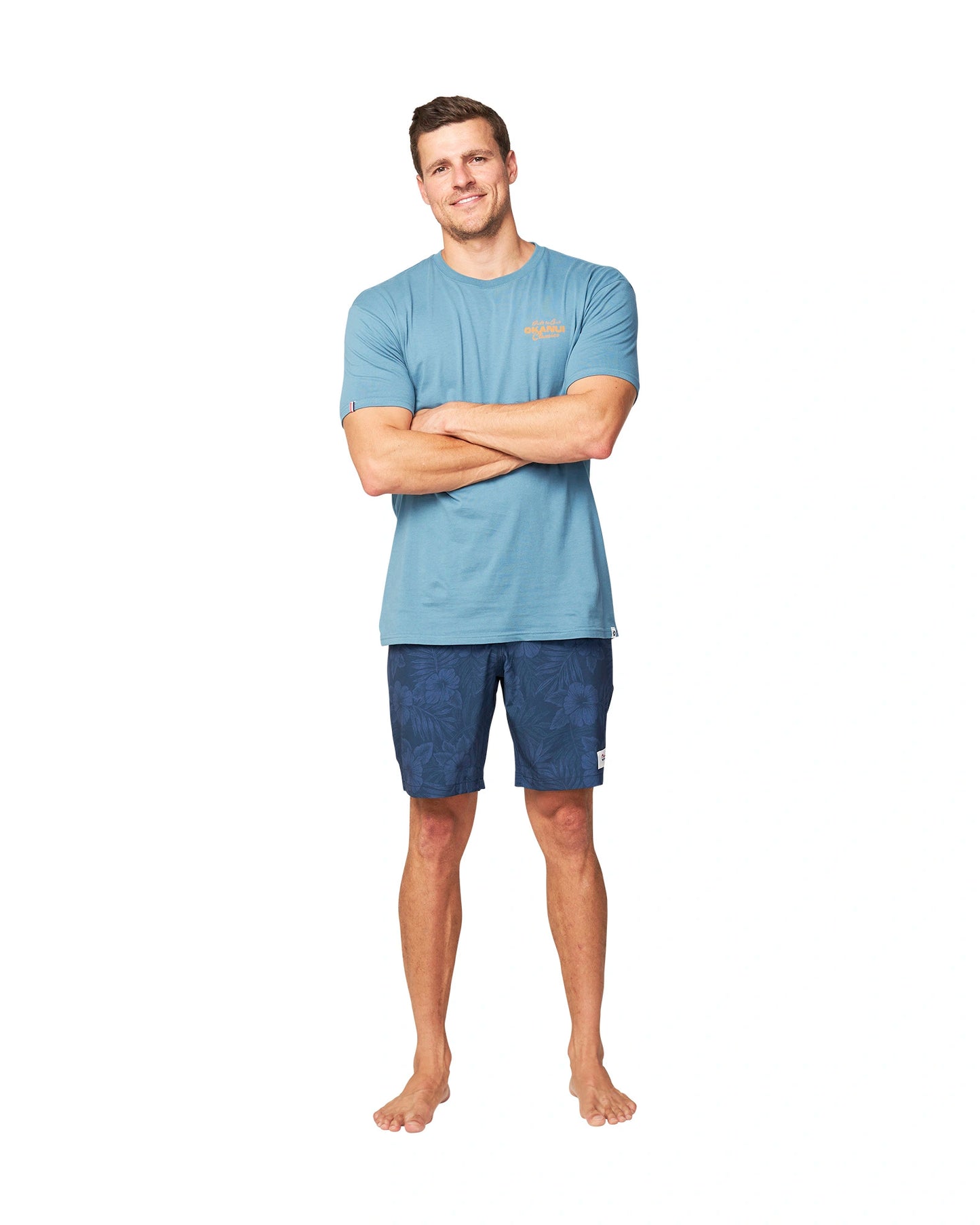 A model wearing a blue t-shirt with his arms crossed and wearing the Okanui Midnight Jungle Boardrider Boardshort in navy color.