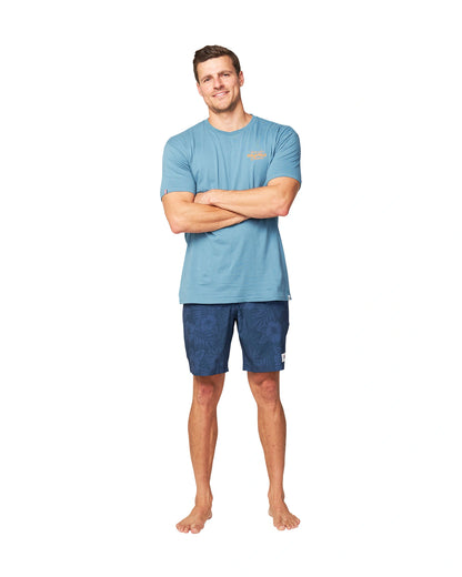 A model wearing a blue t-shirt with his arms crossed and wearing the Okanui Midnight Jungle Boardrider Boardshort in navy color.