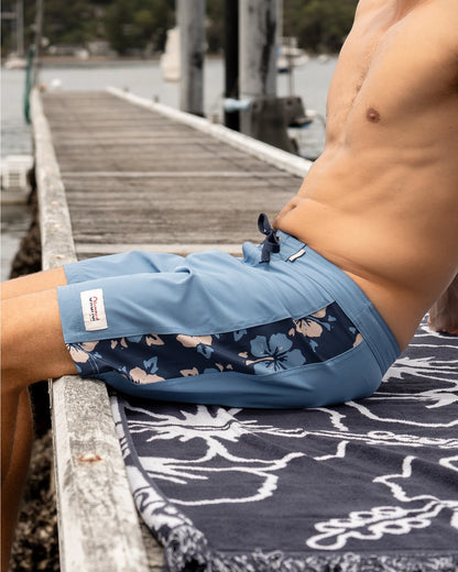 A model sitting on the docks wearing the Okanui Collage Boardrider Boardshort in Steel colour.