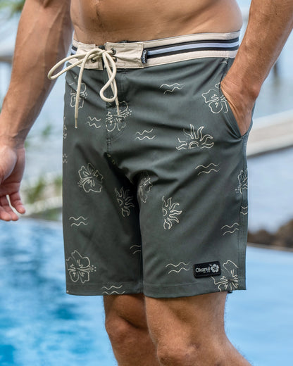 A man standing on the docks with his hand on his pocket wearing the Okanui Rising Sun Boardrider Boardshort in Forest color.