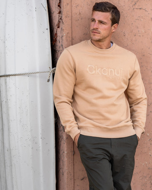 A man dressed in dark pants and a beige Okanui sweatshirt on top. 