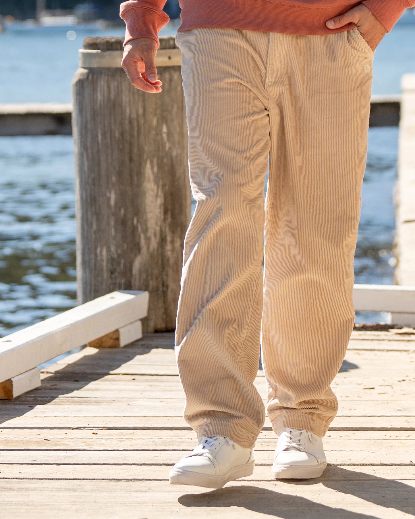 A close up lower view of a walking pose of a male model on the docks wearing the Okanui's Bay Corduroy Pant in Oat colour.