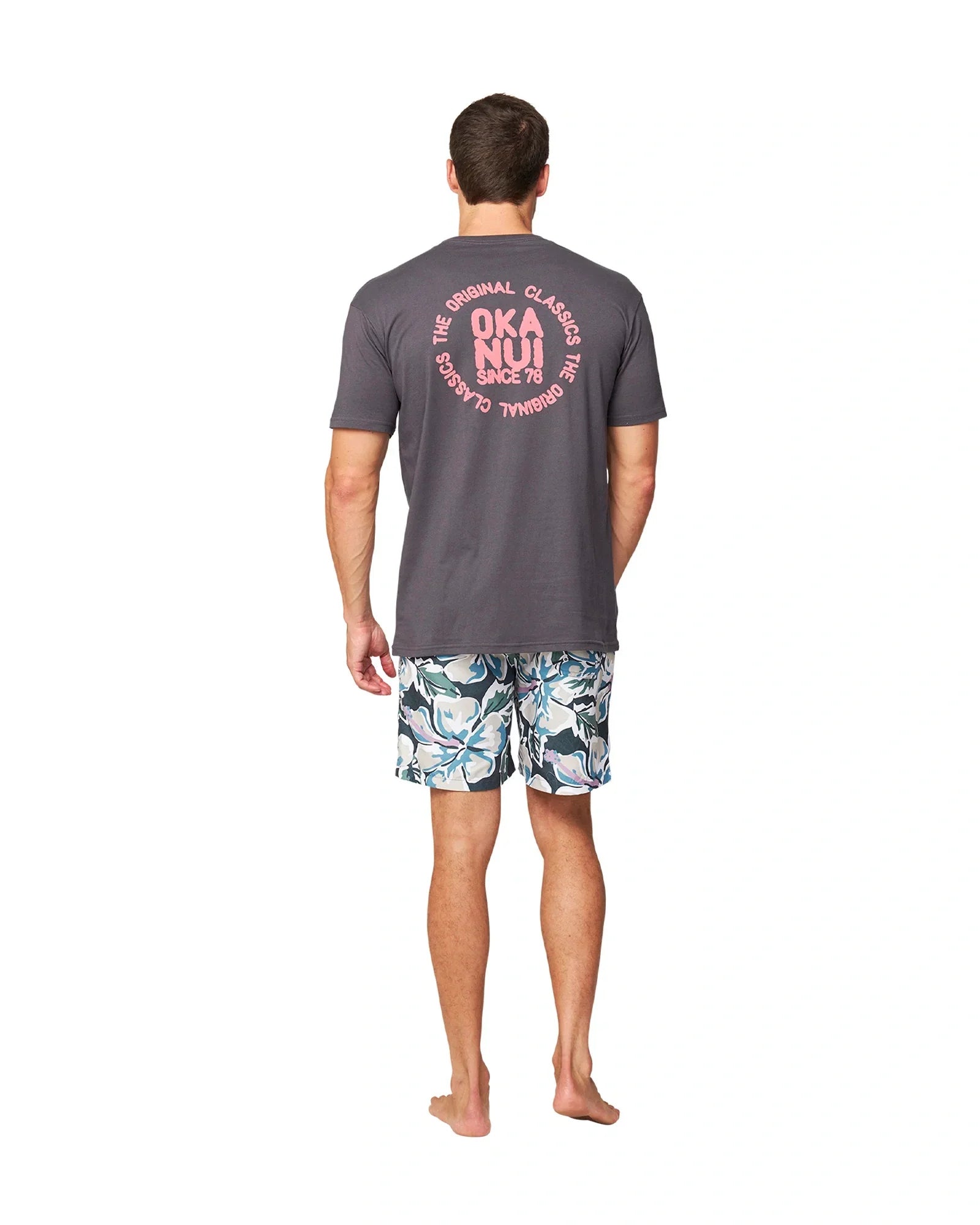 A male model wearing a dark grey t-shirt showcasing the back print with pink font color and the Okanui Flora Fatigue Swim Shorts.