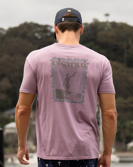 A male model showcasing the back of the Okanui Zine T-Shirt in Washed Lilac colour posing outdoors with trees in the background.