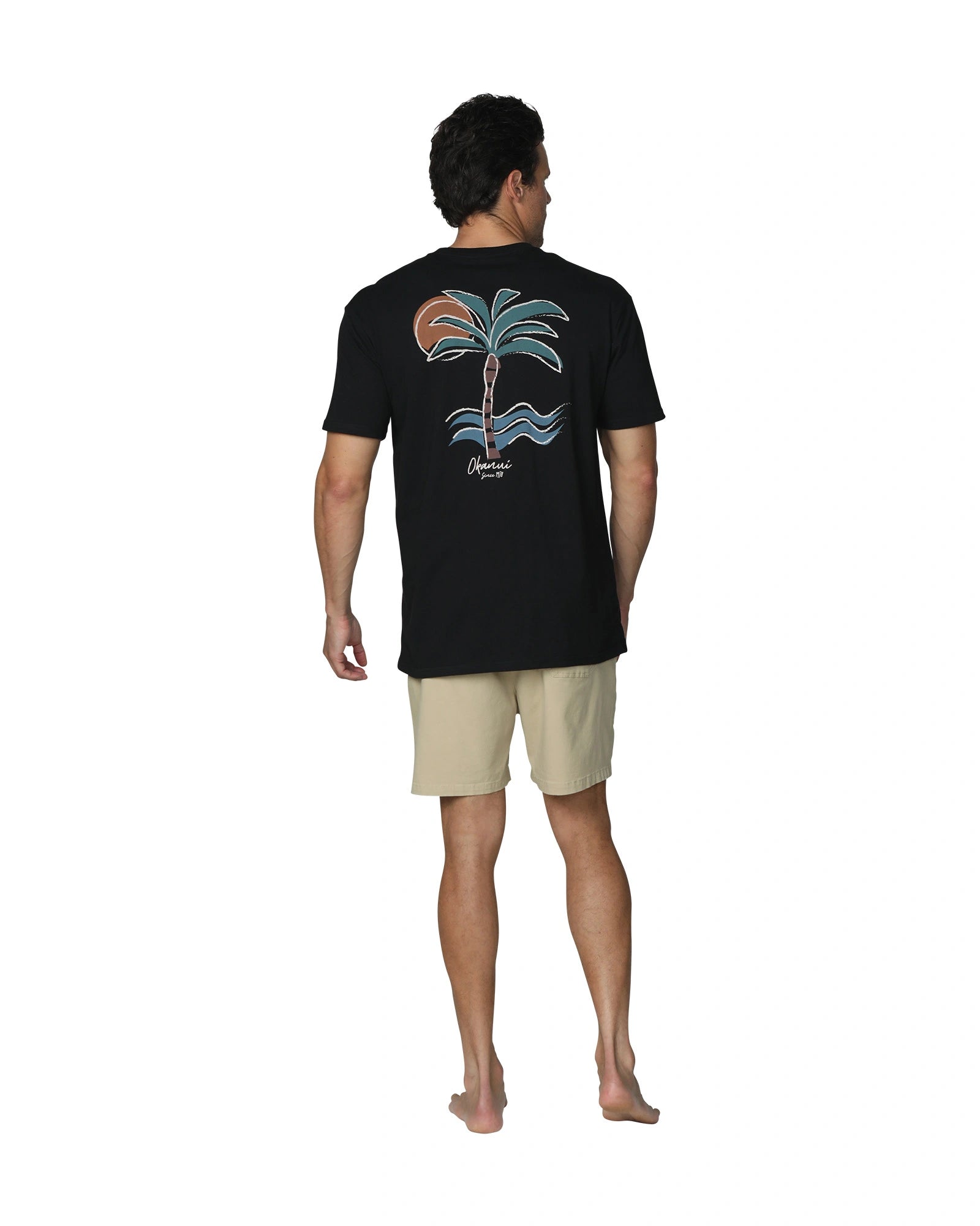 A full back view of a male model wearing the Okanui Island Nights T-Shirt in Black colour featuring a plam tree back print paired with a khaki walkshort.