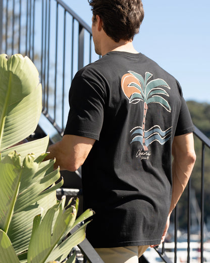 A male model walking up the stairs showcasing the back print of a palm tree of the Okanui's Island Nights T-Shirt in Black colour.