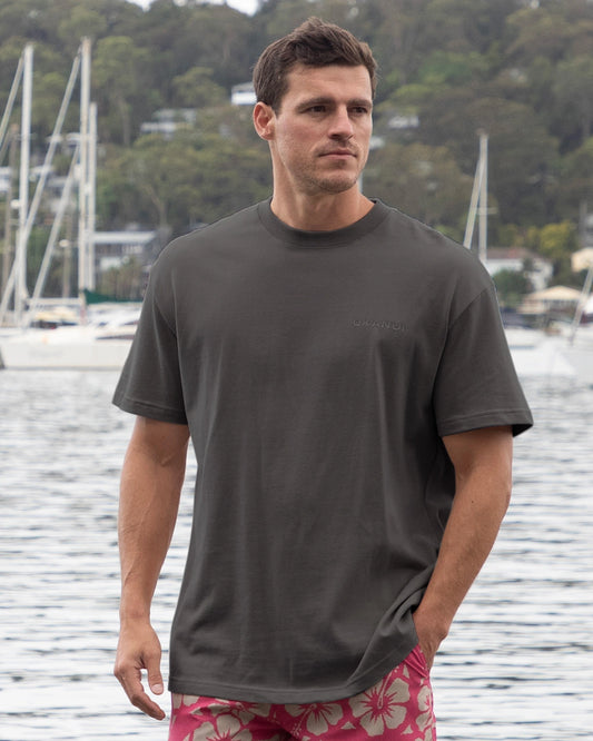 A male model posing oon the docks with boats in the background wearing the Okanui Men's Cube Heavy Weight Tee in Washed Iron colour.