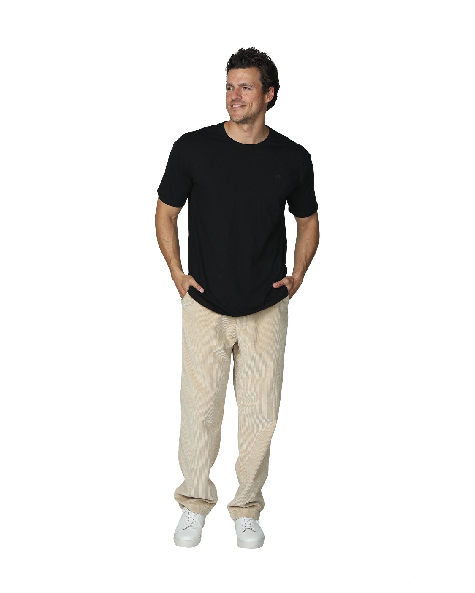 A full body front view of a male model with his hands in his pockets wearing the the Okanui Mens Staple Tee in Black colour paired with a khaki corduroy pants and white shoes.