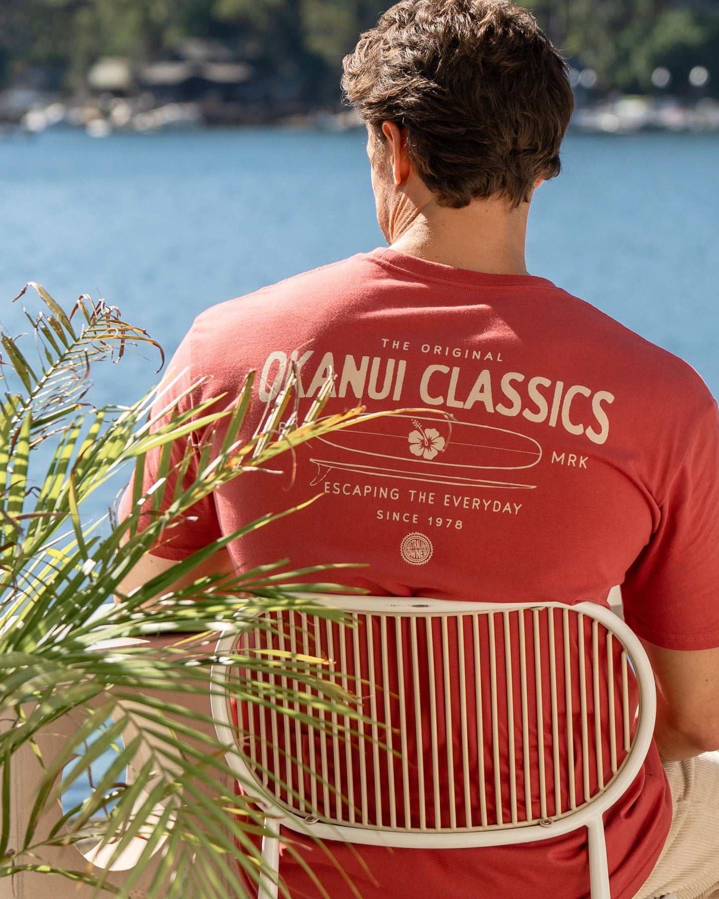 A male model sitting at a porch with a view of the bayside wearing Okanui's Boardriders Men's T-Shirt in Rust colour.