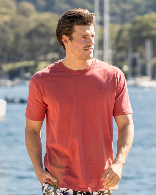 A walking pose of a model at the docks wearing the Okanui Mens Staple Tee in Rust colour with his hand in one pocket.