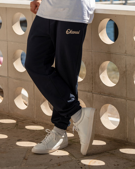 A close up lower view of a male model leaning against a concrete balcony railing wearing the Okanui Navy Signature Track Pants in Navy colour.