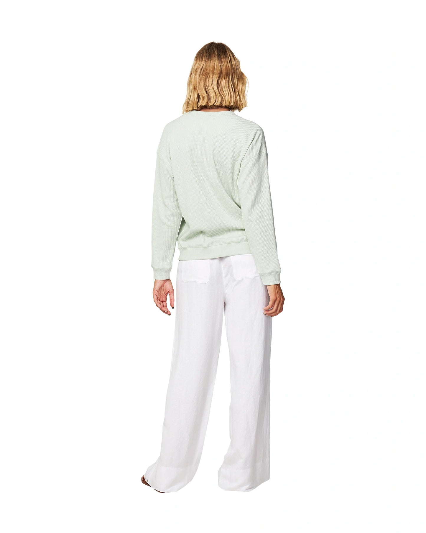 Full body back view of a female model wearing the Okanui long sleeve waffle knit jumper in gelato sage variant paired with a white trouser