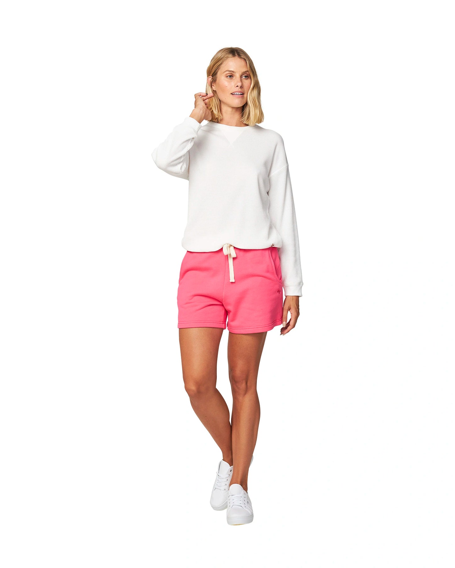 Full body of a female model wearing the Okanui long sleeve waffle knit jumper in white paired with a pink shorts