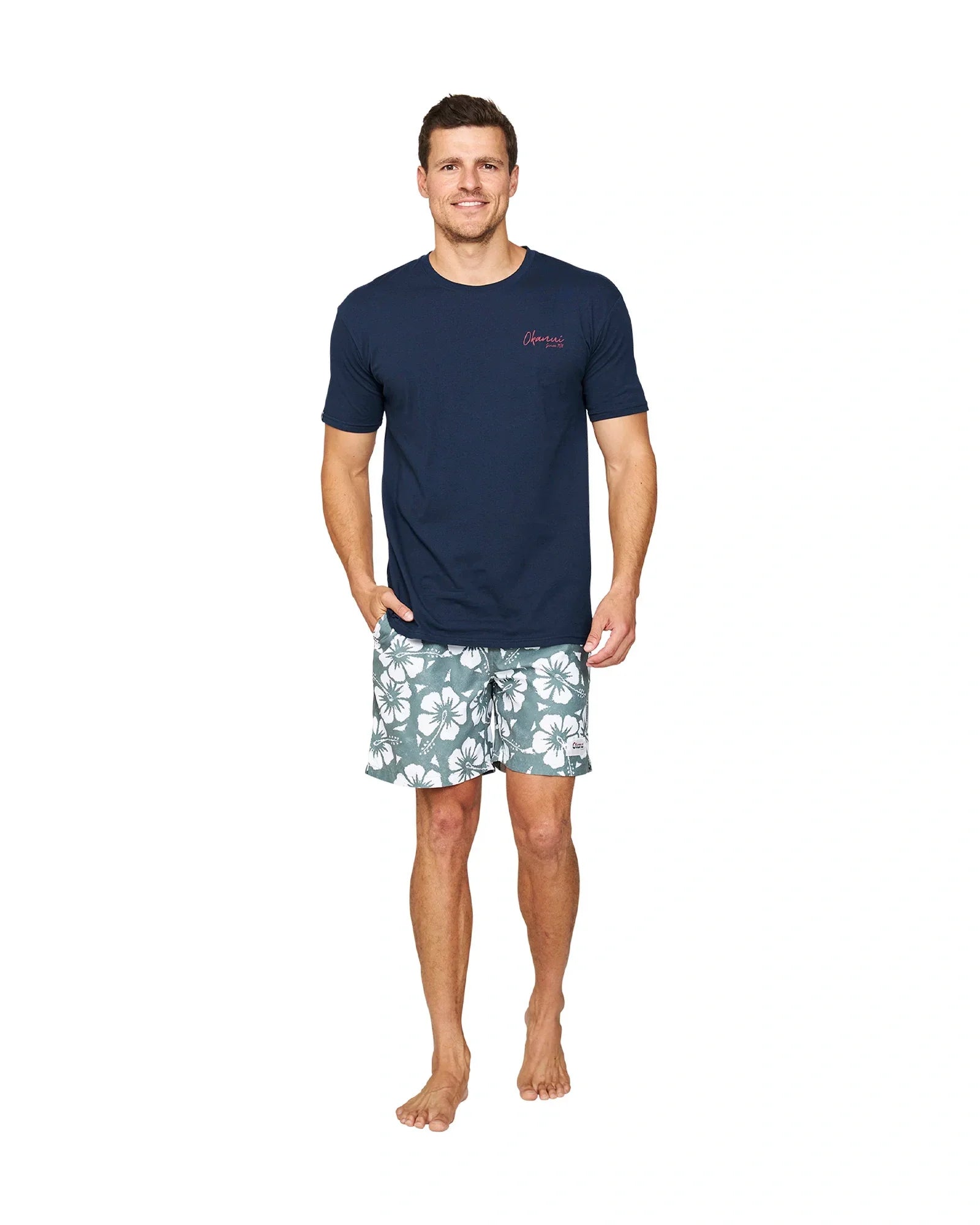 A full body view of a model wearing a navy t-shirt and the Okanui Hibiscus Swimshorts in washed khaki color.