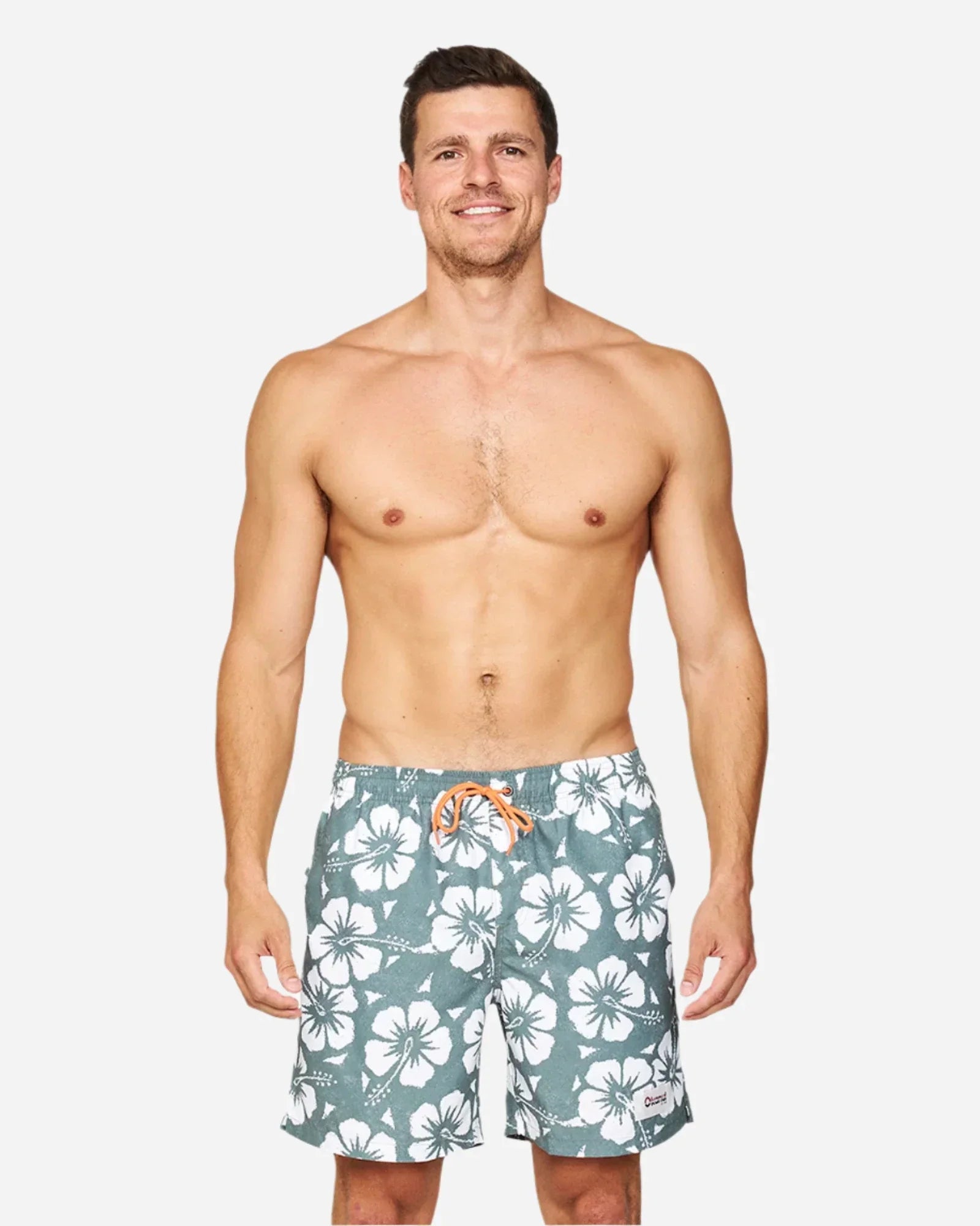 A front view of a smiling male model wearing the Okanui Hibiscus Swimshorts in washed khaki colour.