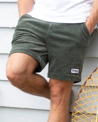 A male model leaning against the wall outside a house wearing a white t-shirt and the Okanui Big Iron Cord Walk Short in Hunter Green colour.