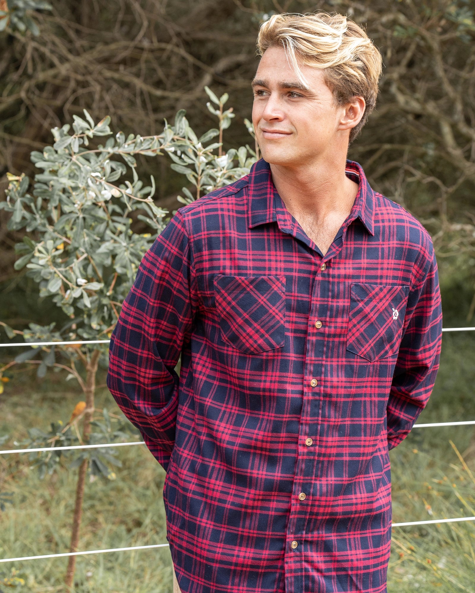 A male model posing candid wearing the red Okanui Flannel Long Sleeve Hiker Shirt with bushes in the background and wire fence.