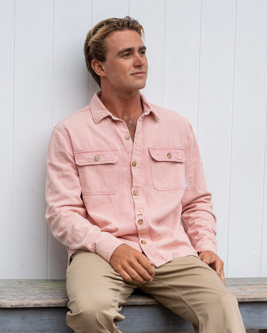 A male model sitting on a bench wearing the Okanui Long Sleeve Deck Shirt in Dusty Pink color.