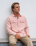 A male model sitting on a bench wearing the Okanui Long Sleeve Deck Shirt in Dusty Pink color.