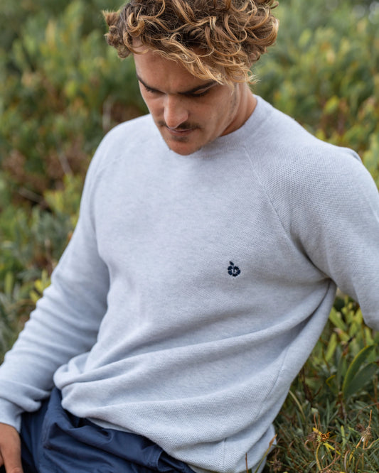 A male model sitting on a bush looking down pose wearing the Okanui Anchor Knit.