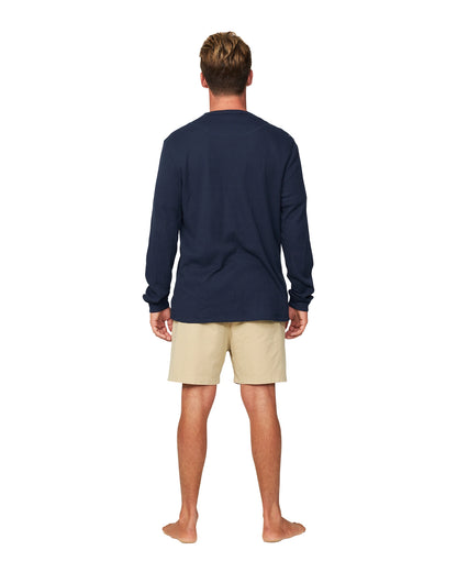 A whole body back view of a male model wearing shorts and the Okanui Long Sleeve Waffle Tee in navy color.