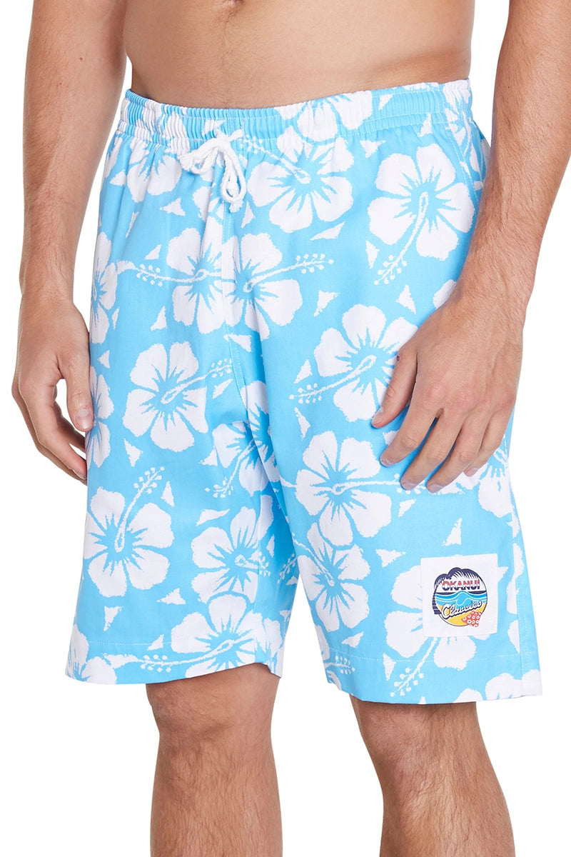 A man is wearing light blue board shorts with color hibiscus skyblue, there ia white flower pattern. The shorts have a white drawstring waist and a patch on the front right leg that features a colorful logo. 