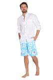 A man with a beard is standing barefoot. He is wearing a white button-up shirt with the sleeves rolled up and light blue shorts adorned with a white floral pattern. His hands are in his pockets, and he faces slightly to the left.