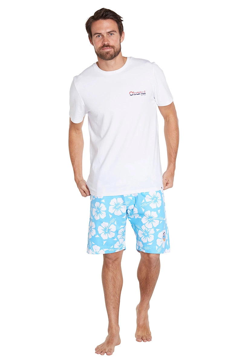 A man with a beard is wearing a white T-shirt and blue floral-patterned shorts. He is barefoot.