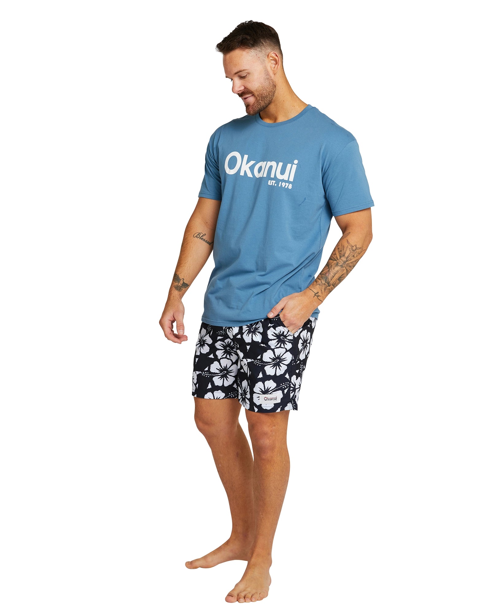 A pose of a male model wearing a blue Okanui t-shirt the Okanui Swimshorts in black colour featuring it's white Hibiscus flower prints.