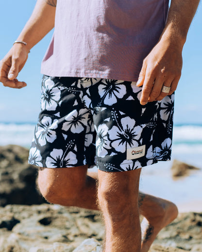 A half body view of a male model at the beach wearing the Okanui Swimshorts in black colour featuring it's white Hibiscus flower prints.