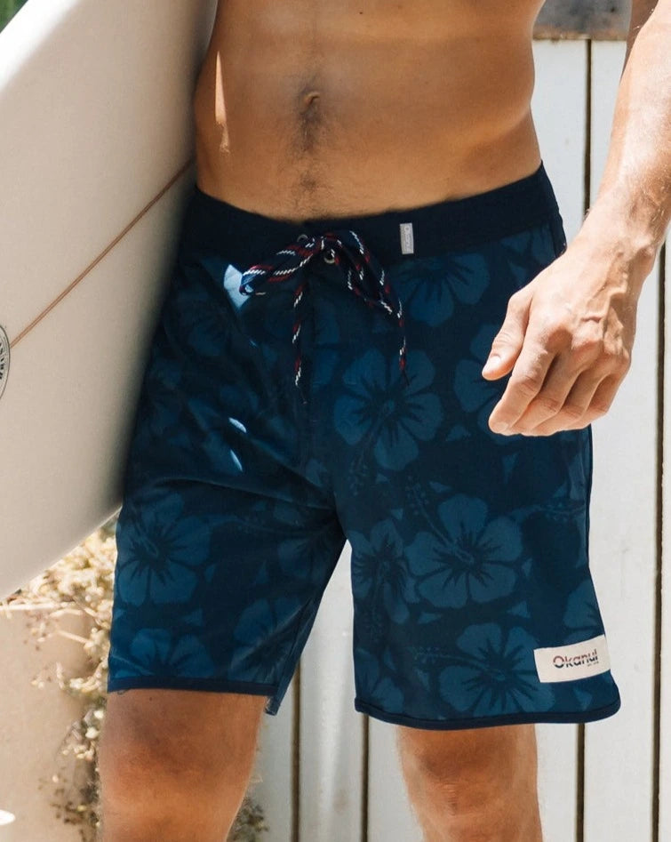 A half body view of a male model wearing the Okanui Boardrider Boardshorts holding a surfboard.