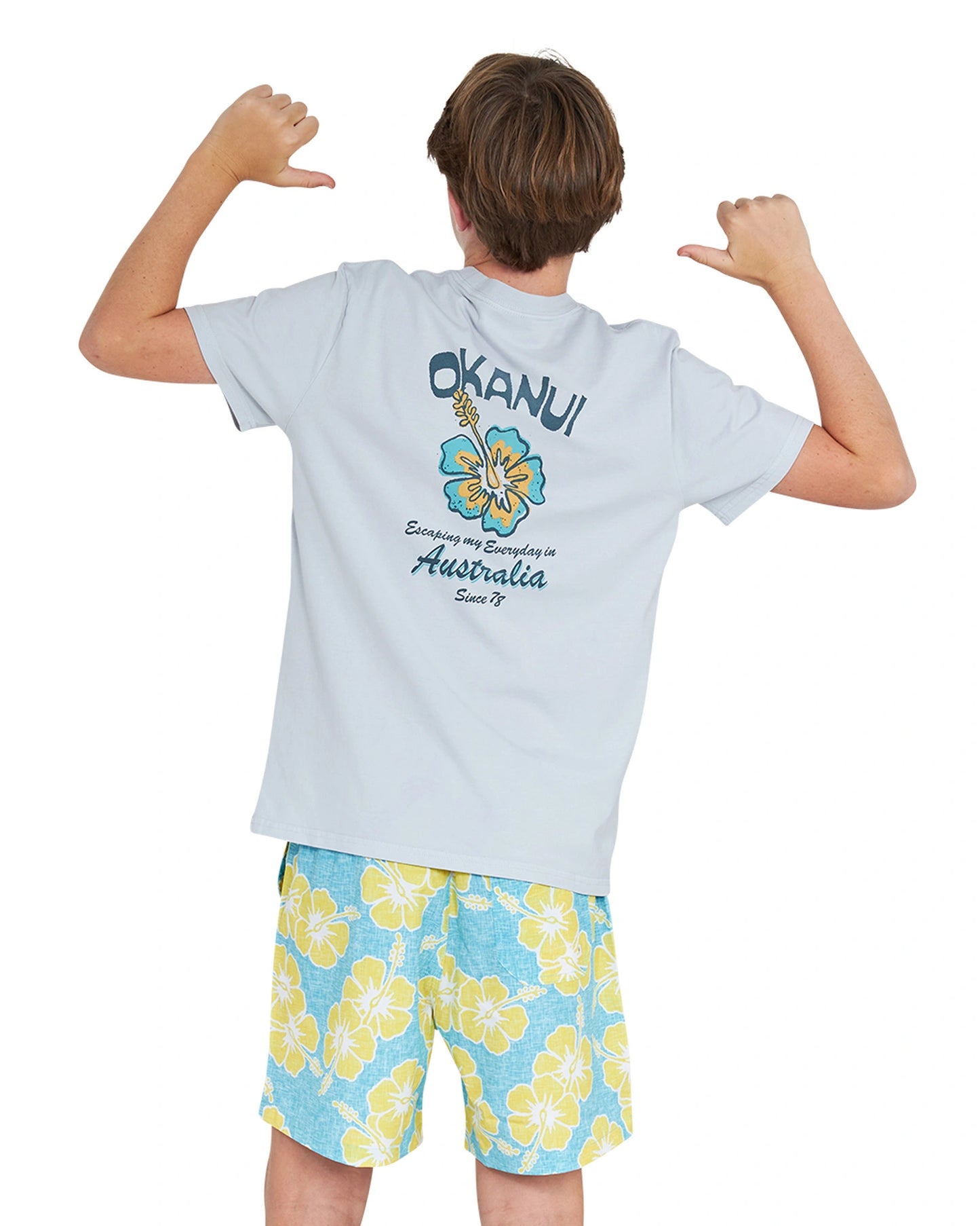 Back view thumbs up pose of a boy wearing the Okanui Way Back When stretch swim short in Aqua yellow paired with white shirt