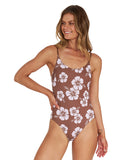A woman wearing a brown hibiscus flower one-piece with her hands on her waist and a smile on her face.