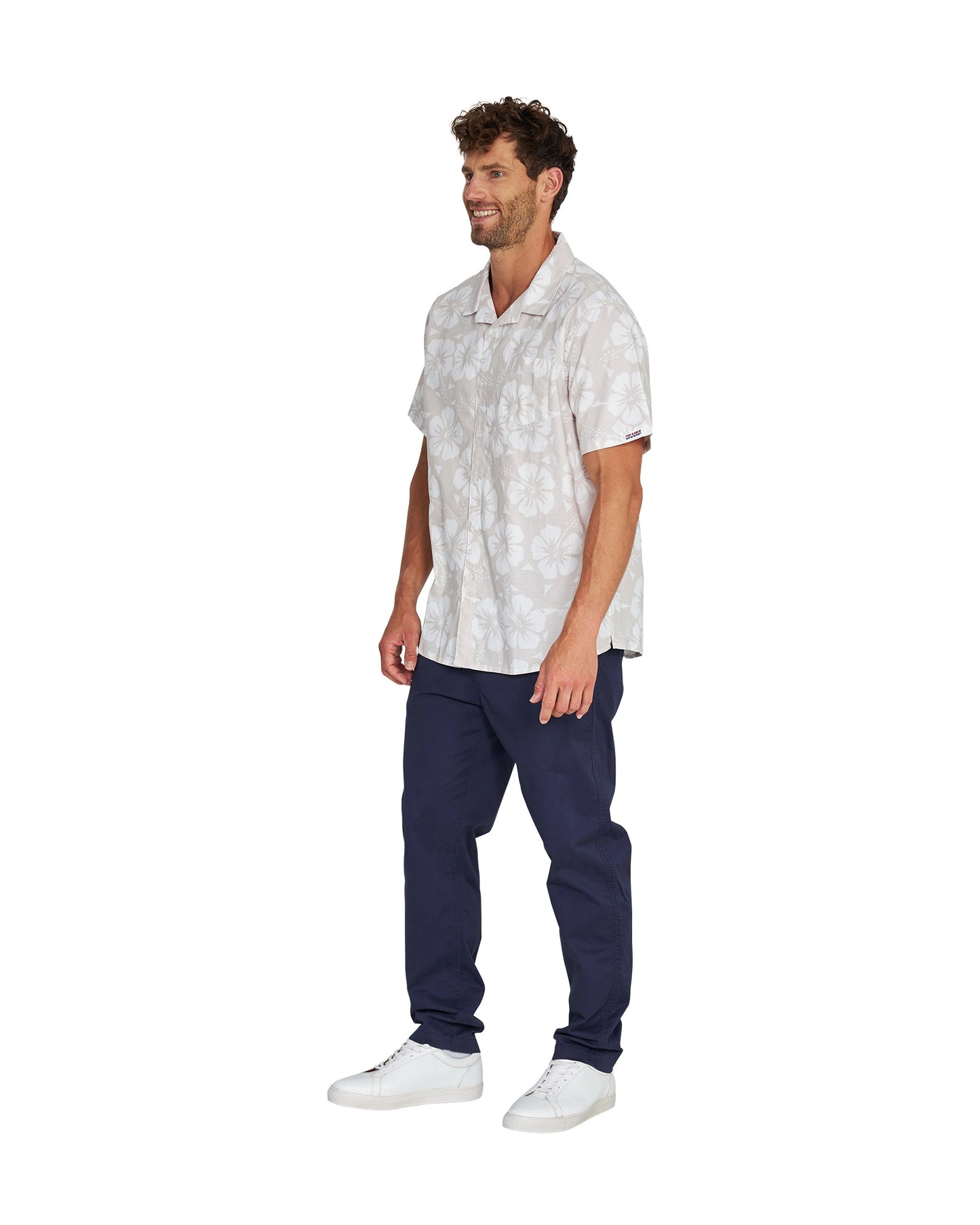 Angled left side view of a male model wearing the Okanui Classic Button up short sleeve shirt in Hibiscus Stone paired wtih a blue pants and white shoes