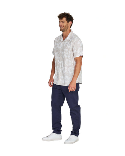 Angled left side view of a male model wearing the Okanui Classic Button up short sleeve shirt in Hibiscus Stone paired wtih a blue pants and white shoes