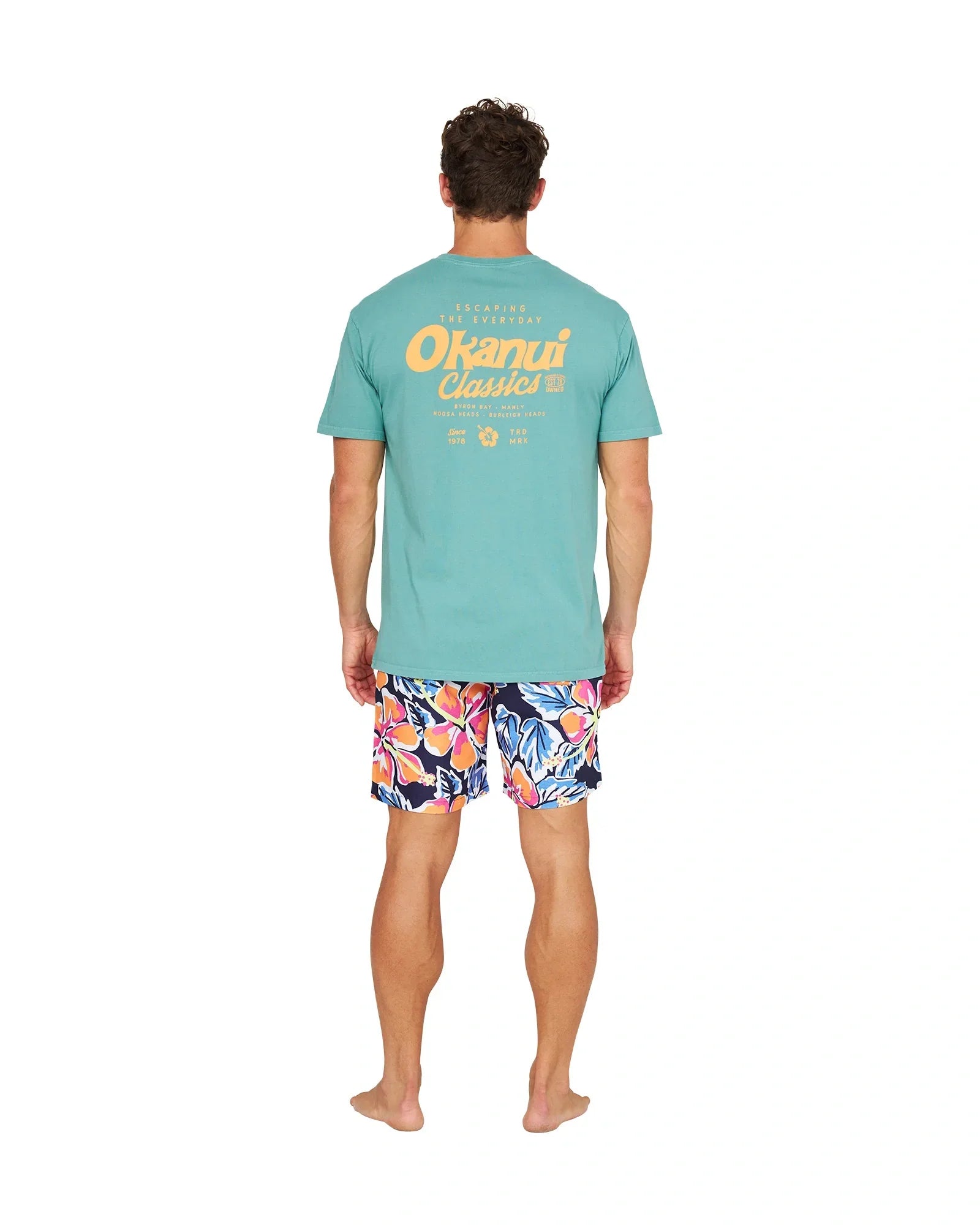 A full body back view of a male model wearing the Okanui Rhapsody Swim Short in navy multi and a light blue shirt with a yellow print of Okanui logo and phrases in yellow font color.