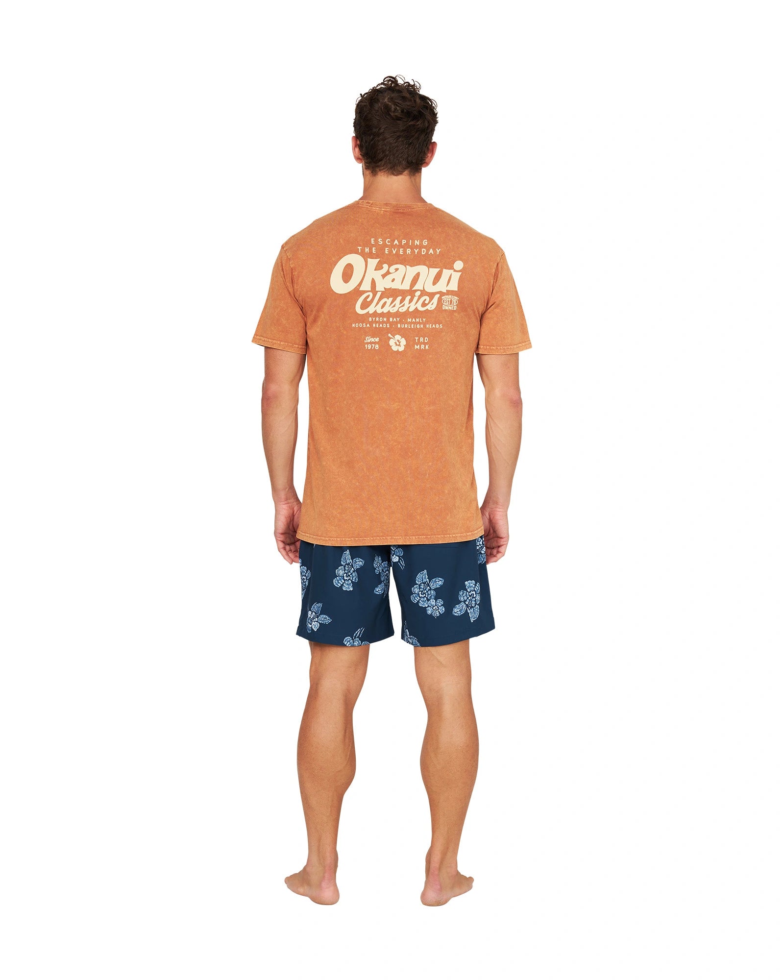 A back view of a male model posing casually wearing the Okanui Big Screen T-Shirt in rust color.